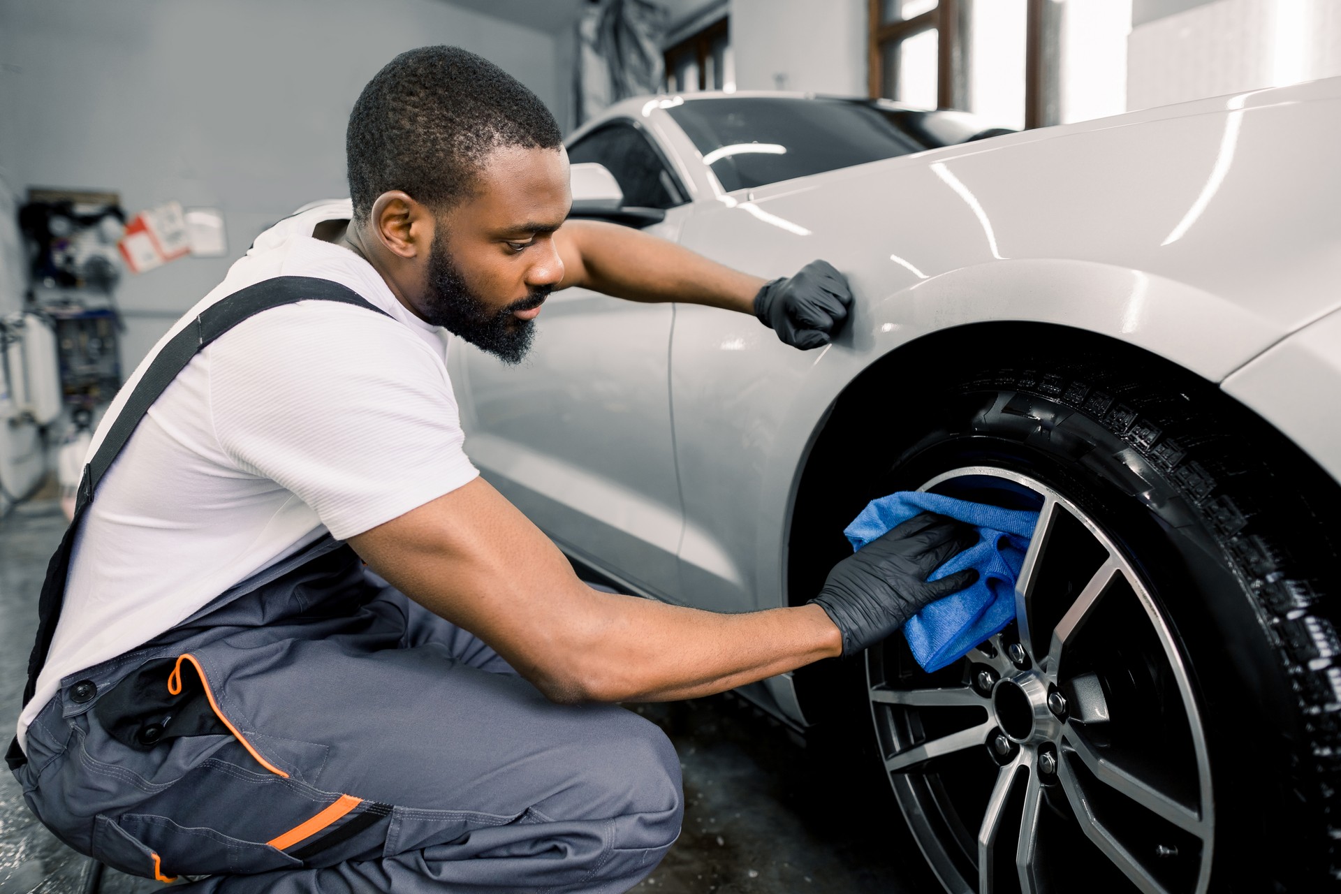 Car washing and detailing photo. African man worker in protective overalls and rubber gloves, washing car alloy wheel on a car wash, using microfiber cloth and special cleaner